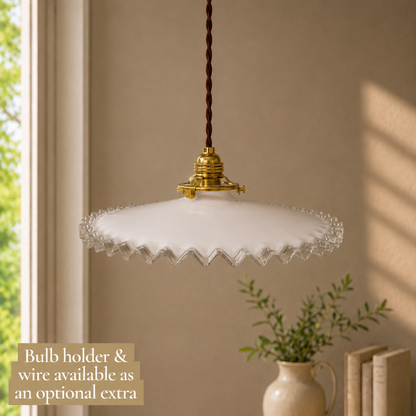 A vintage-style pendant light with a scalloped white glass shade and gold brass fixture, hanging in a warm beige room with a window and a small plant in the background. A text overlay in the bottom left reads 'Bulb holder & wire available as an optional extra'.