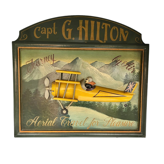 A vintage-style decorative wall sign featuring a painted mountain landscape and a yellow biplane with a small figure on the wing. The sign reads "Capt G Hilton