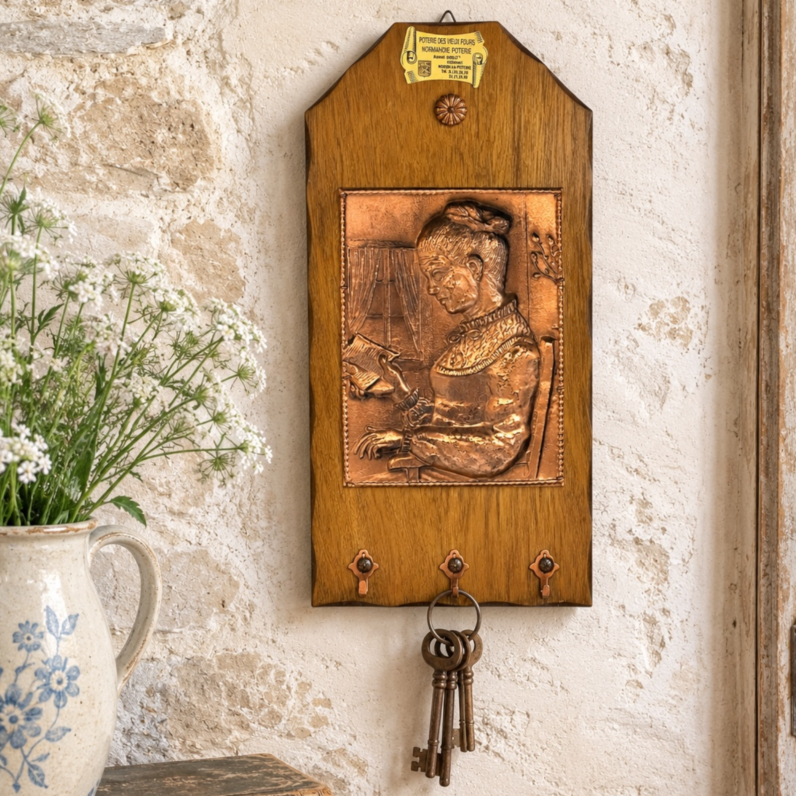 A rustic wooden wall-mounted key holder with a raised copper-toned relief of a woman at a desk, three hanging keys, and a small yellow sign near the top, shown against a textured stone wall with flowers in a ceramic vase at the side.